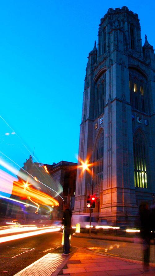 The University of Bristol Law School’s Wills Memorial Building, lit up against a dusky blue sky, with lines of motion captured traffic lights rushing across the foreground.
